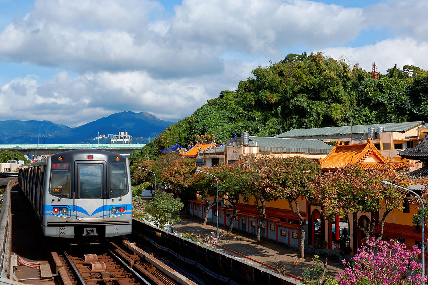 A metro Train traveling on elevated rails of Taipei MRT System by the architecture of a Buddhist temple near Yuanshan station with mountains in background on a sunny cloudy day in Taipei, Taiwan; Shutterstock ID 794476882; purchase_order: -; job: -; client: -; other: -